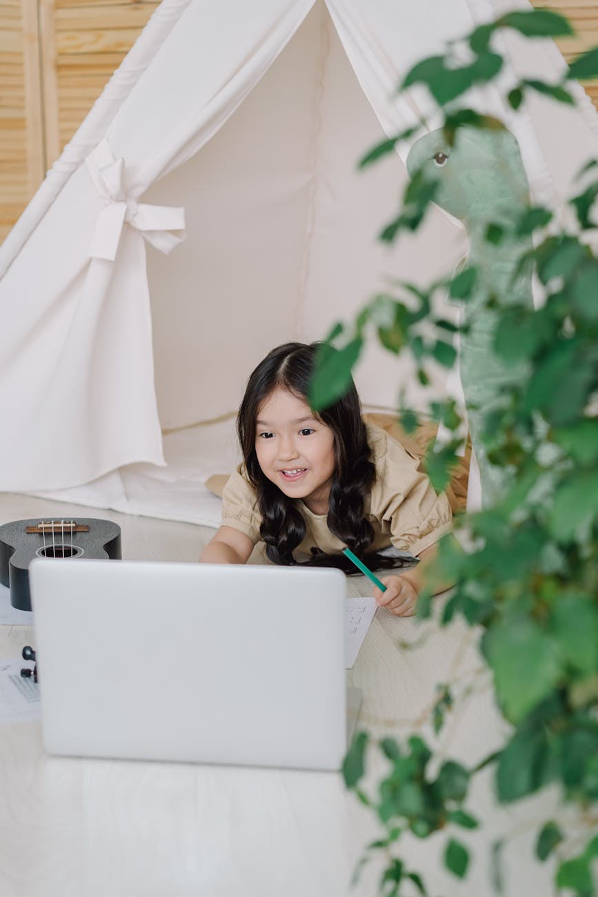 A young girl smiles while interacting with a laptop inside a cozy indoor tent, surrounded by a relaxing home setting.